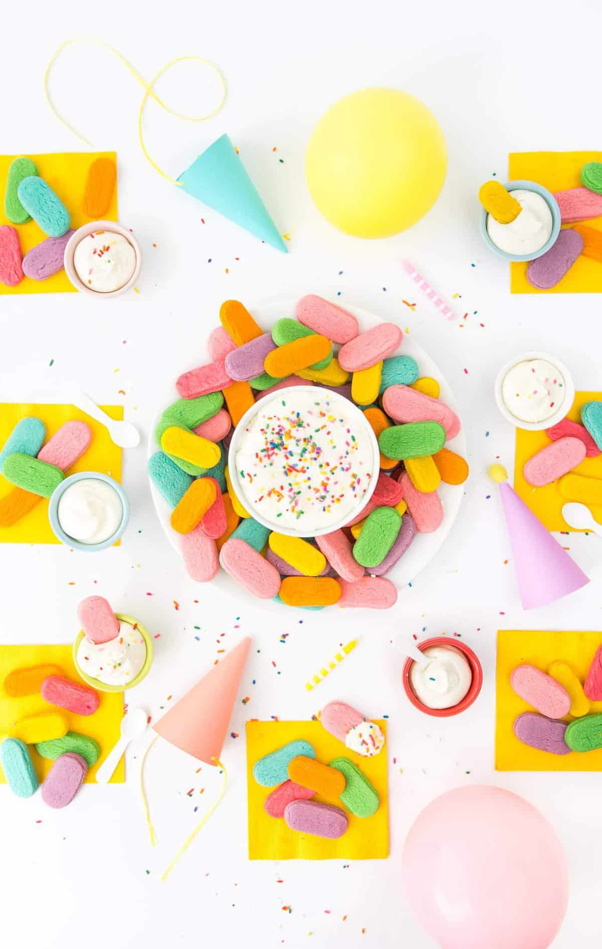 Overhead photo of party table with plate of rainbow sugar cookies on yellow napkins, balloons, and paper party hats.