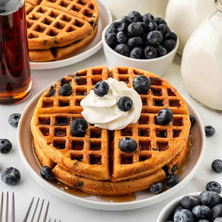 Stack of blueberry waffles with whipped cream on top with blueberries.