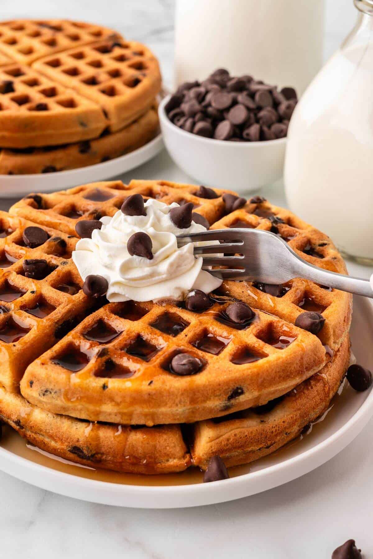Fork cutting into stack of chocolate chip waffles with whipped cream.