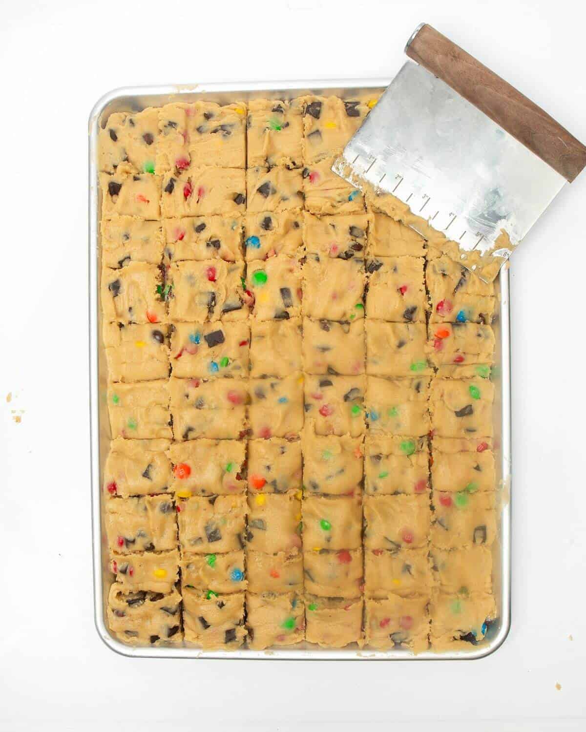 Using bench scraper to cut cookie dough into squares in metal pan.