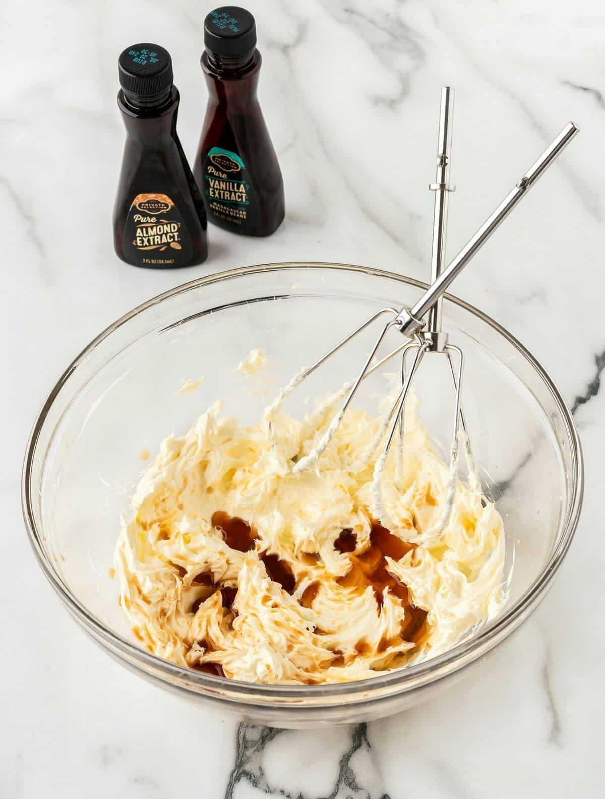 buttercream frosting in clear mixing bowl with two beaters and almond and vanilla extract