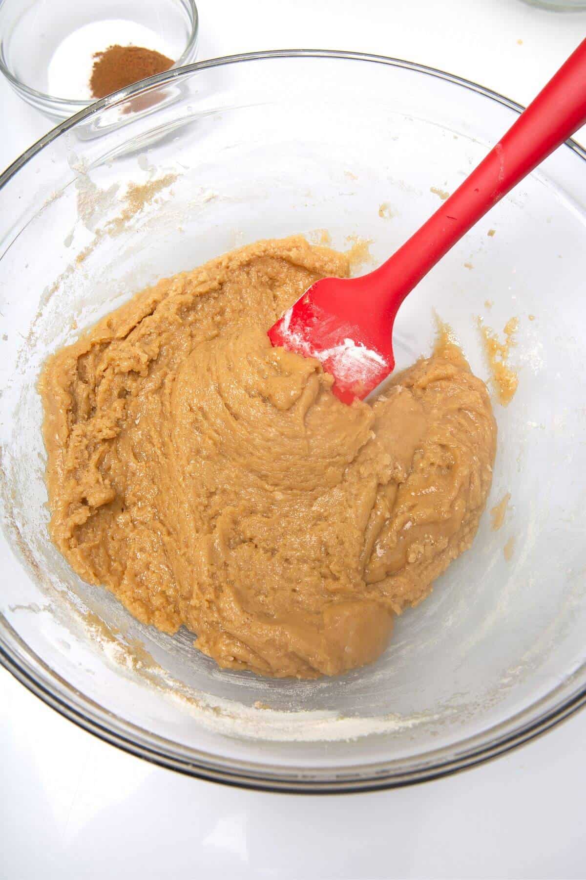 Snickerdoodle cookie cake dough in glass mixing bowl and red spatula.