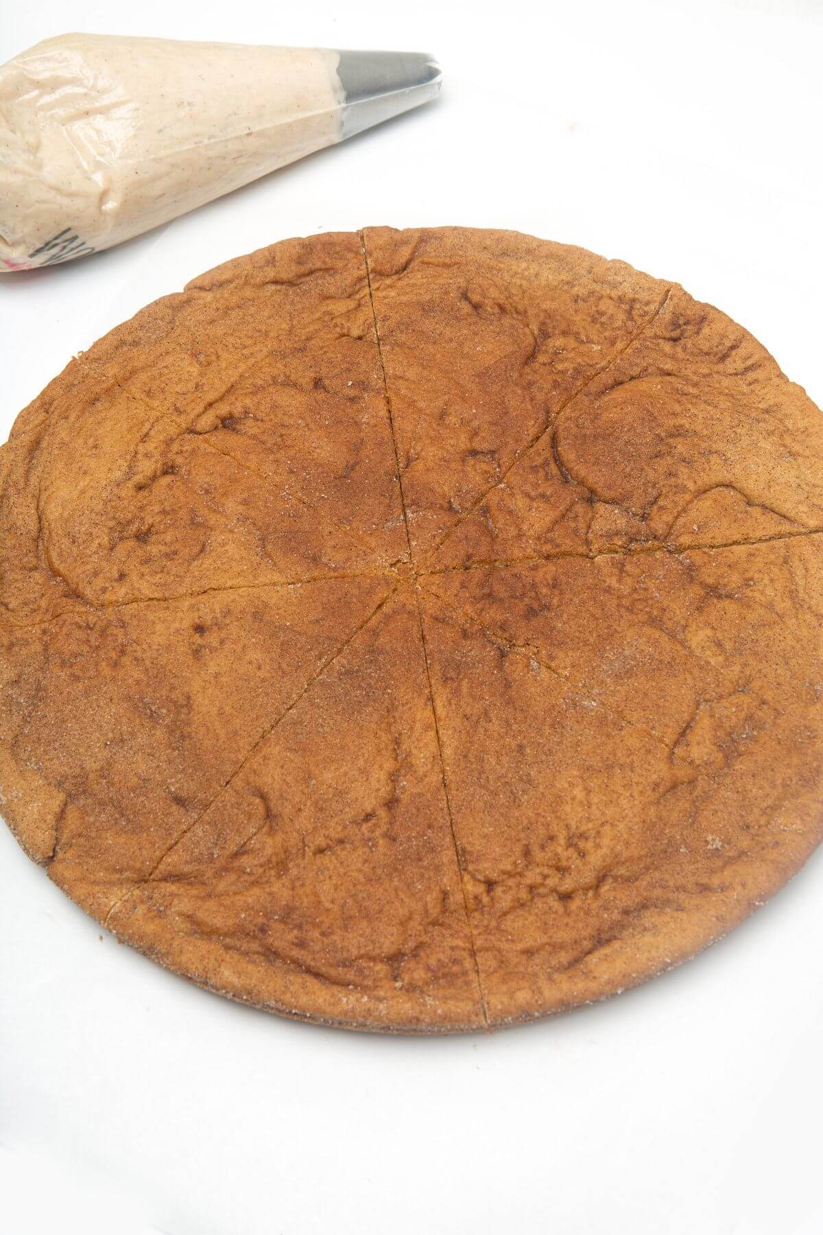 Baked snickerdoodle cookie cake with bag of frosting behind it.